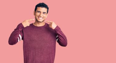 Gündelik kıyafetler giyen, neşeli gülümseyen, parmaklarıyla ve ağzıyla işaret eden İspanyol asıllı yakışıklı genç adam. diş sağlığı kavramı. 