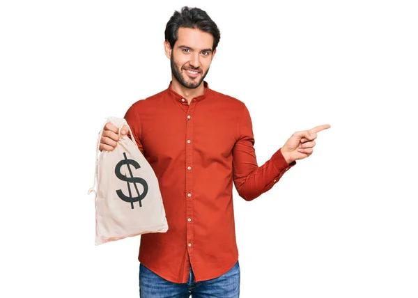 Joven Hispano Sosteniendo Bolsa Dólares Sonriendo Feliz Señalando Con Mano —  Fotos de Stock