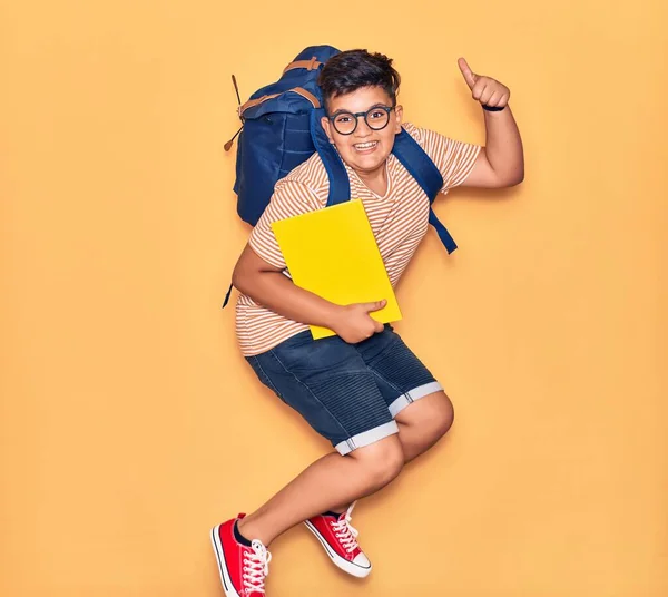 Adorable student boy wearing glasses and backpack smiling happy. Jumping with smile on face holding book doing ok sign with thumb up over isolated yellow background.