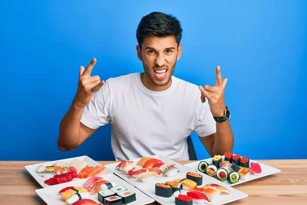 Young handsome man eating sushi sitting on the table shouting with crazy expression doing rock symbol with hands up. music star. heavy concept.