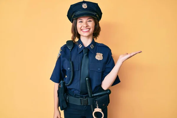 Young beautiful woman wearing police uniform smiling cheerful presenting and pointing with palm of hand looking at the camera.
