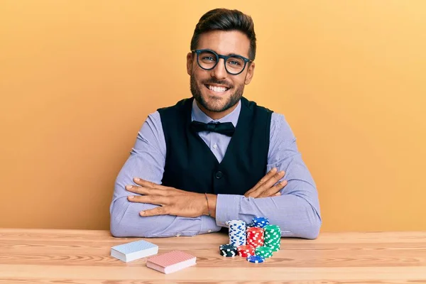 英俊而惊慌失措的男人坐在桌子上 拿着扑克牌和扑克牌开心地微笑着 交叉着双臂看着相机 积极的人 — 图库照片