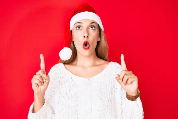 若いですブルネット女性身に着けているクリスマスの帽子驚きと驚きを見上げと指摘とともに指と上げ腕 — ストック写真