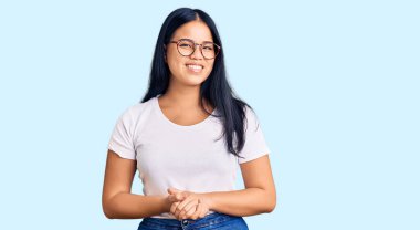 Genç, güzel Asyalı bir kız, günlük kıyafetler ve gözlük takıyor ve ellerini kavuşturup gülümsüyor. Rahatlamış ve neşeli. Başarı ve iyimserlik 