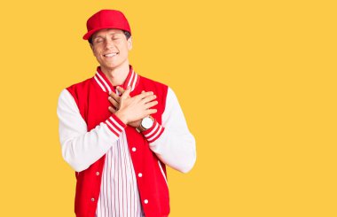 Beyzbol üniforması giyen, elleri göğsünde, gözleri kapalı gülümseyen ve yüzünde minnet ifadesi olan genç yakışıklı bir adam. sağlık kavramı. 