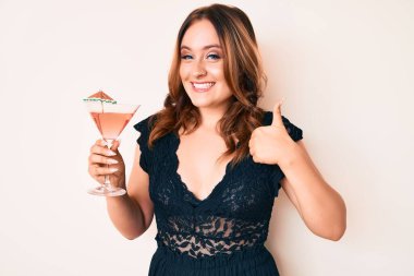 Genç, beyaz bir kadın alkolle tropikal kokteyl içiyor mutlu ve pozitif gülümsüyor, başparmağıyla mükemmel ve onay işareti yapıyor. 