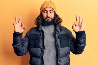 Gündelik kışlık kıyafetler giyen genç Arap adam rahatla ve parmaklarıyla meditasyon hareketi yaparken gözleri kapalı gülümse. yoga konsepti. 