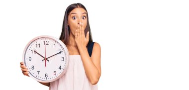 Genç İspanyol bir kadın, büyük bir saat tutuyor. Ağzını eliyle kapatıyor. Şok geçiriyor ve hata yapmaktan korkuyor. şaşırmış ifade 