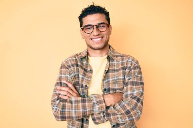 Sıradan kıyafetler ve gözlük takan, kollarını kavuşturup kameraya gülümseyen genç, yakışıklı İspanyol adam. pozitif kişi. 