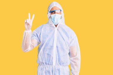 Doktor korumalı Coronavirus üniforması giyen genç İspanyol adam ve tıbbi maske iki numarayı gösterip gösterirken kendinden emin ve mutlu bir şekilde gülümsüyor.. 