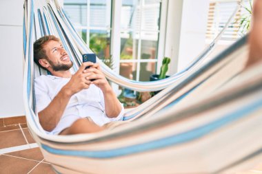 Yakışıklı beyaz adam mutlu bir şekilde gülümsüyor terasta dinlenirken akıllı telefon kullanıyor.