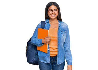 Sırt çantası takan ve elinde kitaplarla gülümseyen genç bir Latin kız, doğal bir ifade. Kendinden emin gülen. 