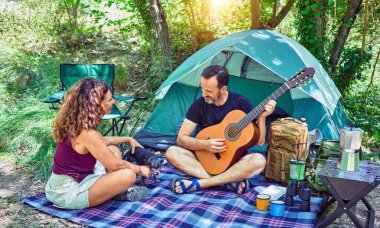 Ormanda kamp yaparken gülümseyen orta yaşlı bir çift. Yerde oturup klasik gitar çalarak şarkı söylemek.