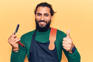 Önlük takan genç Arap adam elinde berber jileti, gülümsüyor mutlu ve pozitif, başparmak yukarı ve onay işareti yapıyor. 