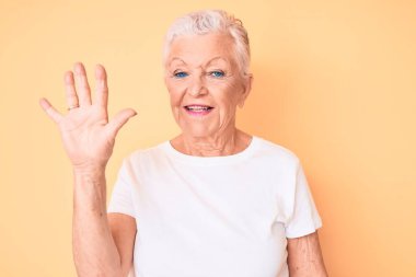 Son sınıf güzeli, mavi gözlü, gri saçlı, sarı arka planda klasik beyaz tişört giyen ve beş numarayı parmaklarıyla gösteren, kendinden emin ve mutlu gülümseyen bir kadın.. 