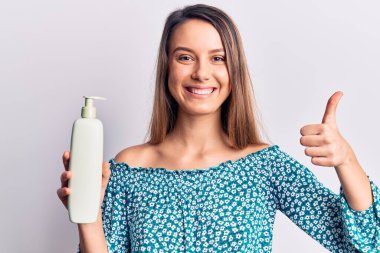 Genç, güzel bir kız elinde krem losyonu şişesiyle mutlu ve pozitif gülümsüyor, başparmağıyla mükemmel ve onay işareti yapıyor. 