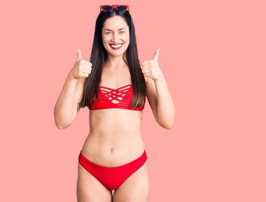 Genç, beyaz, güzel bir kadın bikini giyer. Başarı işareti yapar. Elleriyle olumlu jest yapar. Neşeli ifade ve kazanan jest. 