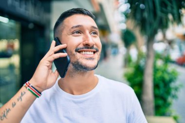 Genç İspanyol gülümseyen adam şehrin caddesinde akıllı telefondan mutlu bir şekilde konuşuyor..