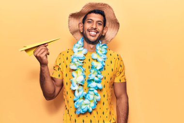 Yaz şapkası takan genç bir latin adam ve elinde kağıt uçak tutan Hawaii zambağı. Pozitif ve mutlu görünüyor. Dişlerini gösteren bir gülümsemeyle ayakta duruyor. 