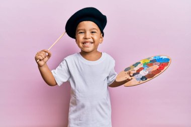 Sevimli Latin çocuk gülümsüyor. Ressam tarzı giyiniyor. Boya fırçası ve palet kullanıyor..