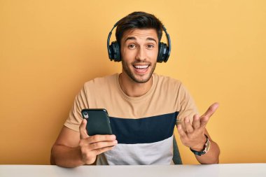 Akıllı telefon takıp başarıyı kutlamak için kulaklık takan genç ve yakışıklı bir adam mutlu bir gülümsemeyle ve elini kaldırarak kazanan bir ifade takınıyor. 