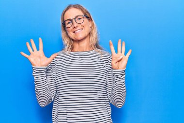 Orta yaşlı, beyaz, çizgili süveter ve gözlük giyen sarışın kadın dokuz numaralı parmaklarını gösterip gösterirken kendine güvenen ve mutlu bir şekilde gülümsüyor.. 