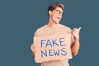 Elinde sahte haber afişi tutan yakışıklı genç adam başparmağını yana kaldırıyor ve ağzı açık gülümsüyor. 