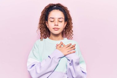 Kıvırcık saçlı, sıradan spor kıyafetler giyen, göğsünde elleri, yüzünde minnet dolu bir jestle gülümseyen güzel bir kız çocuğu. sağlık kavramı. 