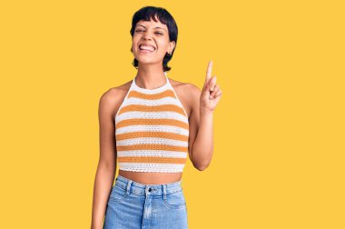 Kısa saçlı genç esmer kadın günlük giysiler giyiyor. Bir numarayı işaret ediyor ve gülümsüyor. Kendinden emin ve mutlu.. 