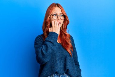 Genç, güzel, kızıl saçlı bir kadın mavi arka plan üzerinde gözlük ve süveter takıyor. Elleri ağız yiyen tırnaklarla stresli ve gergin görünüyor. anksiyete. 