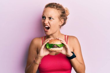 Güzel, beyaz bir kadın lezzetli klasik bir burger yiyor kızgın ve öfkeli çığlıklar atıyor öfkeyle bağırıyor. öfke ve agresif konsept. 