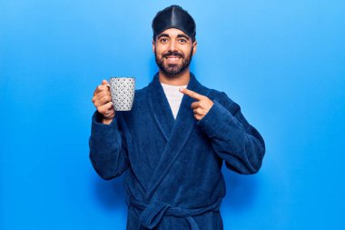 Uyku maskesi ve bornoz takan genç İspanyol adam kahve içiyor mutlu bir şekilde el ve parmak işareti yapıyor. 