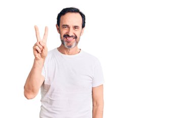 Orta yaşlı yakışıklı bir adam sıradan tişörtler giyer ve parmaklarıyla iki numarayı gösterirken kendinden emin ve mutlu bir şekilde gülümser.. 
