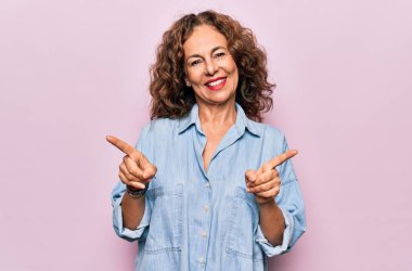Orta yaşlı, güzel bir kadın pembe arka planda dikilen rahat bir kot gömlek giyiyor. Kendine güvenen, farklı yönleri gösteren bir gülümseme. Reklam için boşluk kopyala