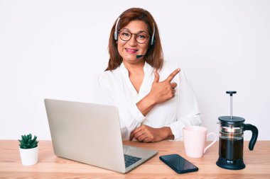 Orta yaşlı esmer, ofiste çalışan, operatör kulaklığı takan, elleri ve parmakları yana doğru gülümseyen bir kadın. 