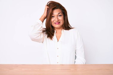 Orta yaşlı esmer, Latin iş kadını, gündelik beyaz gömleğini giymiş masanın üzerinde oturuyor ve soru sormayı merak ediyor. Şüpheyle kararsız, kafa kafaya düşünerek. Düşünceli kavram. 