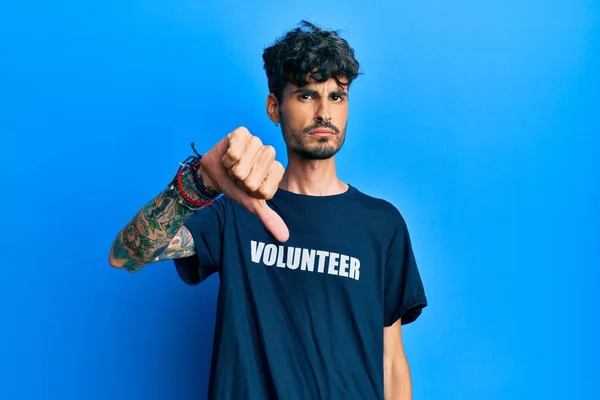 ボランティアTシャツを着た若いヒスパニック系の男は 親指を下にジェスチャーで拒絶と否定を示す不満と怒りを見ています 悪い表現 — ストック写真