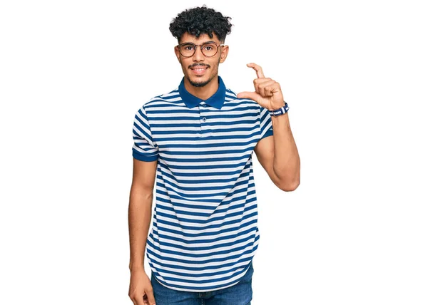 年轻的阿拉伯男子 身穿休闲装 戴着眼镜 微笑着 自信地做手势 手拿着小尺寸的标志 手拿着手指 手拿着相机 计量概念 — 图库照片