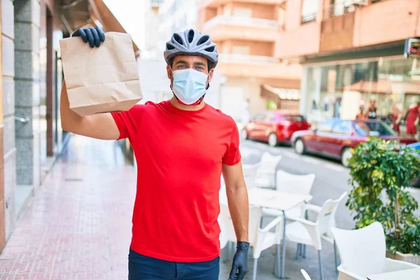 若い配達人は自転車のヘルメットとコロナウイルス保護医療用マスクを着て街の通りで紙袋を奪う — ストック写真