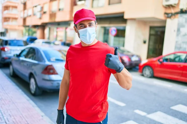街の通りに立って制服やコロナウイルス対策医療マスクを身に着けている若い配達人 — ストック写真