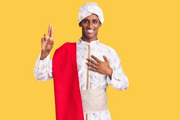 African handsome man wearing tradition sherwani saree clothes smiling swearing with hand on chest and fingers up, making a loyalty promise oath