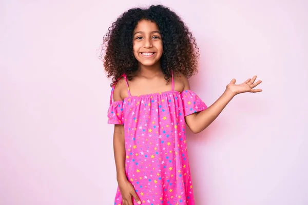 カジュアルなドレスを身に着けている巻き毛を持つアフリカ系アメリカ人の子供は陽気なプレゼンテーション笑顔とカメラを見て手の手のひらで指す — ストック写真