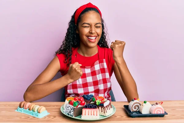 Młoda Afroamerykanka Mundurze Piekarza Siedząca Stole Słodyczami Świętującymi Sukces Podniesionymi — Zdjęcie stockowe
