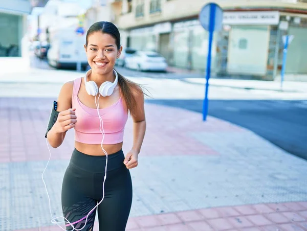 若い美しいヒスパニックスポーツの女性は ランナーの服とヘッドフォンを着て 町で走る幸せ笑顔 — ストック写真