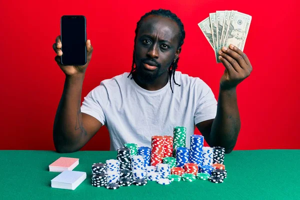 衝撃的な顔でスマートフォンやドルを保持ポーカーをプレイハンサムな若い黒人男性 懐疑的で皮肉を探して オープン口で驚いて — ストック写真