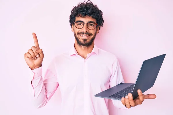 Handsome young man with curly hair and bear working using computer laptop surprised with an idea or question pointing finger with happy face, number one