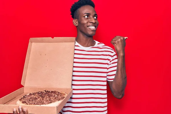 年轻的非洲裔美国人拿着送披萨盒 用大拇指指向旁边 笑着张开了嘴 — 图库照片