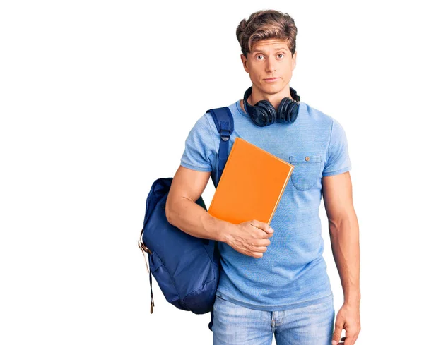 年轻英俊的男子 背着学生背包和耳机 持着书本思维的态度和严肃的表情 看上去很自信 — 图库照片