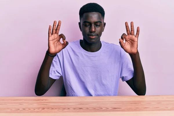 若いアフリカ系アメリカ人の男性は テーブルの上に座ってカジュアルな服を着てリラックスし 指で瞑想のジェスチャーを行う目を閉じて笑顔 ヨガのコンセプト — ストック写真