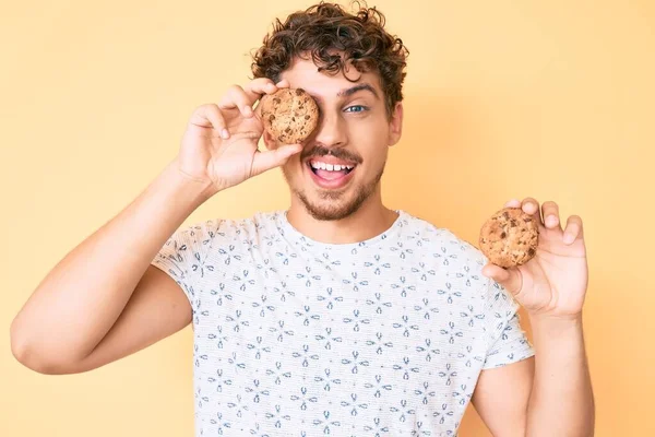 Young caucasian man with curly hair holding cookie winking looking at the camera with sexy expression, cheerful and happy face. — Stock Image Young Caucasian Man Curly Hair Holding Cookie Winking Looking Camera Stock Picture